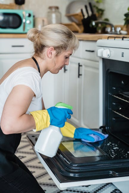A woman wearing yellow and blue cleaning gloves is performing surface cleaning on the interior of a black oven in a kitchen. The kitchen features white cabinetry with black handles, a wooden counterto