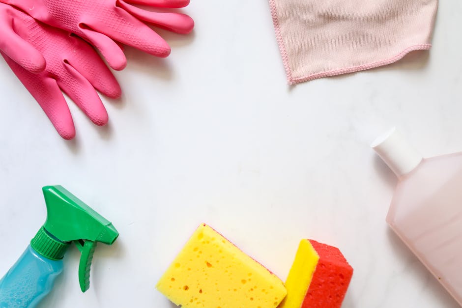 An arrangement of cleaning supplies on a white surface, including a pair of pink rubber gloves at the top left, a pink cloth at the top right, a blue spray bottle with a green trigger at the bottom le